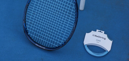 tennis racket on a hard court next to restring zero tennis strings