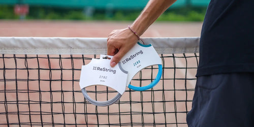 man holding shaped string and round tennis string leaning against tennis net